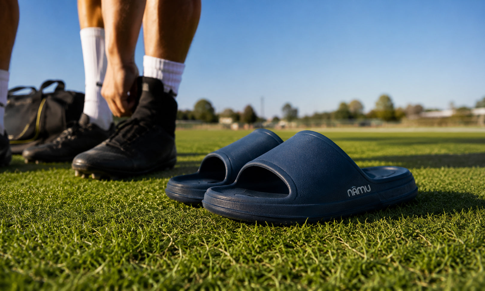 Person wearing black sports shoes and blue slides on a grassy field with a clear sky.